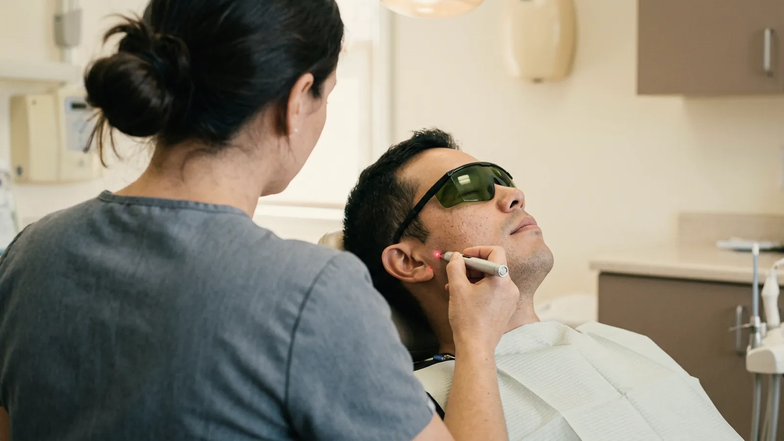 Dentista de costas aplicando laser de baixa intensidade na articulação da mandíbula de um paciente de óculos de proteção